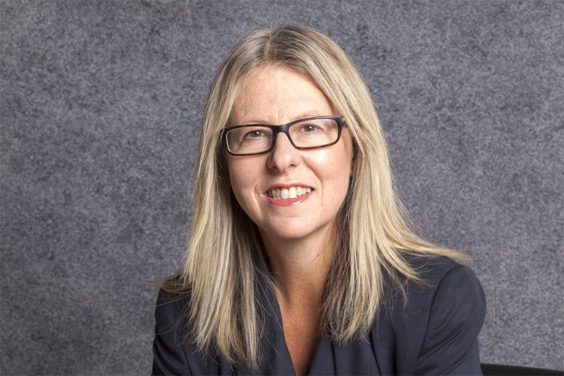 Brenda Crossman smiling, in a dark grey blazer, glasses with straight blond hair down.