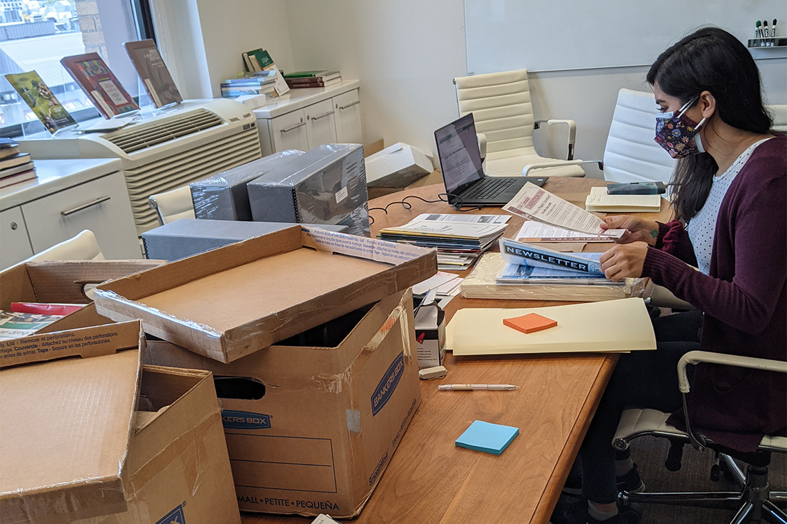 Young South Asian woman sits at a large table surrounded by archival materials, file boxes and office equipment.