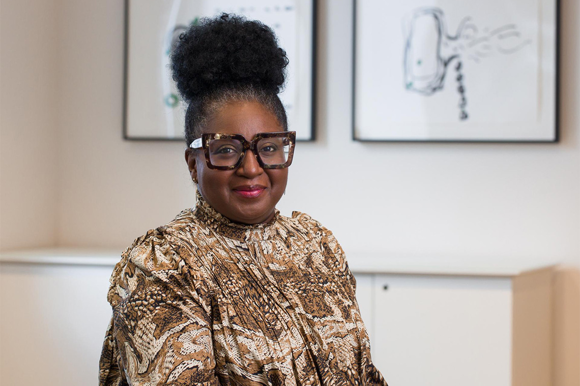 Photo of a smiling Black woman with a bun, wearing brown glasses, a brown-and-white patterned blouse